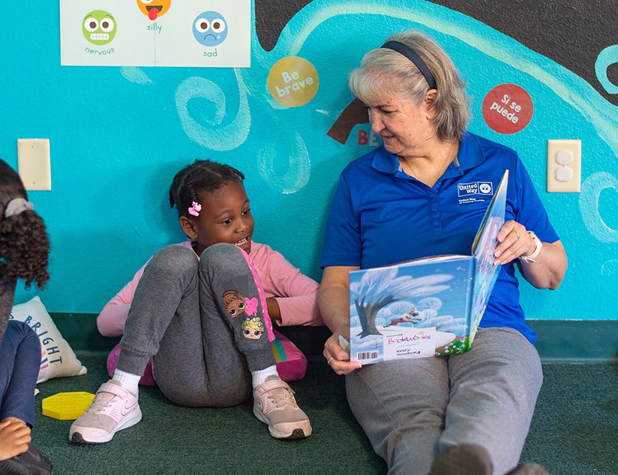 Pam Armstrong wears a blue shirt and sits on the floor next to a 4 year old girl, reading her a book.
