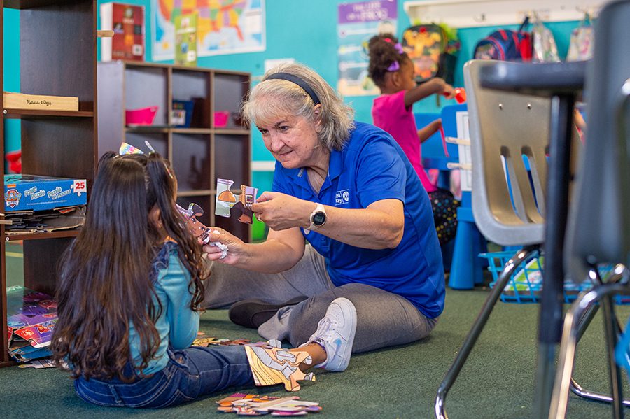 Pam Armstrong sits on the floor holding puzzle pieces up to a young girl, helping her choose her next piece.